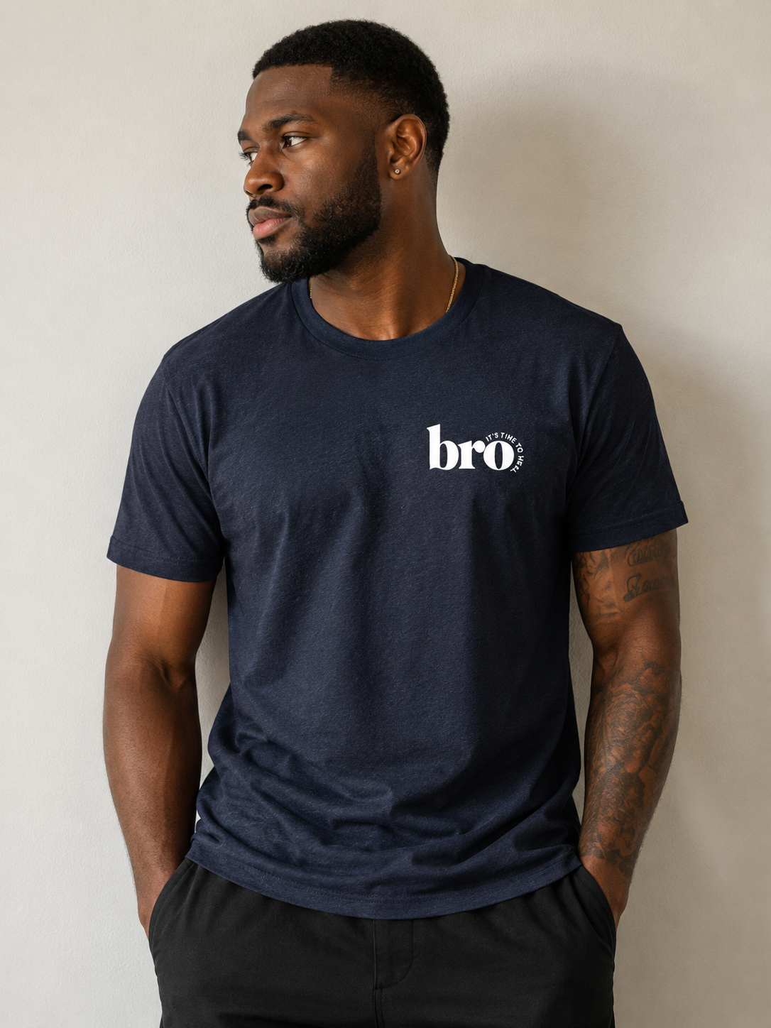 A man wearing a navy blue crewneck t-shirt with a white 'bro' graphic on the left chest, paired with black pants, standing against a neutral beige wall.