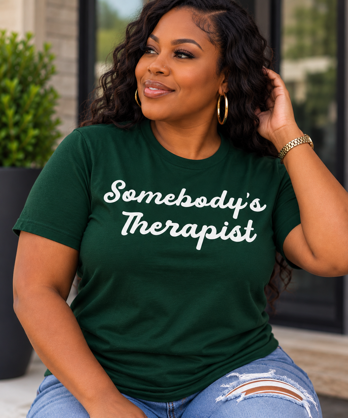 A woman wearing a dark green t-shirt with the phrase 'Somebody's Therapist' printed in white script across the chest, paired with distressed blue jeans.