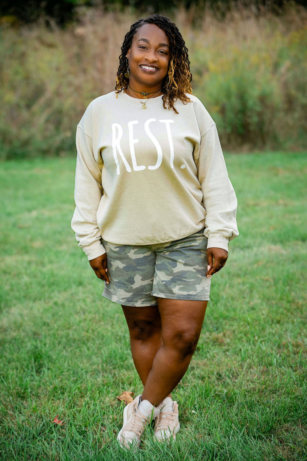 Woman wearing a beige sweatshirt with 'REST' and camouflage shorts standing in a grassy field.
