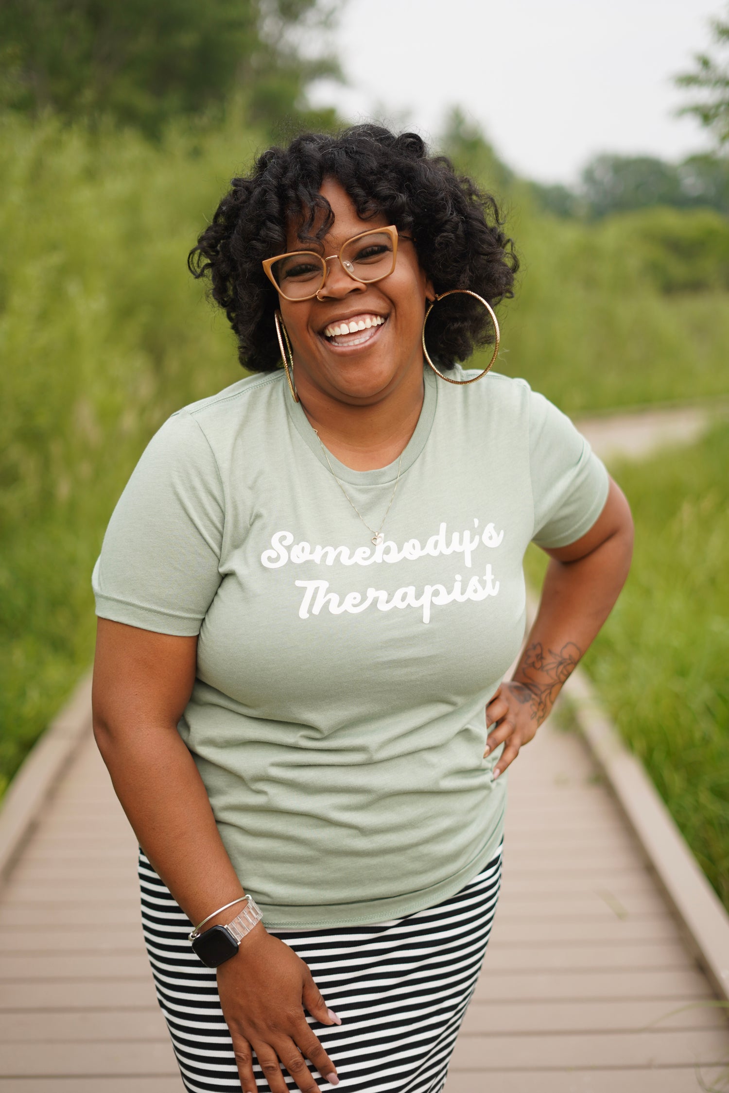 A woman smiling on a wooden boardwalk outdoors, wearing a sage green graphic t-shirt that reads 'Somebody's Therapist' in white script, paired with black and white striped pants.