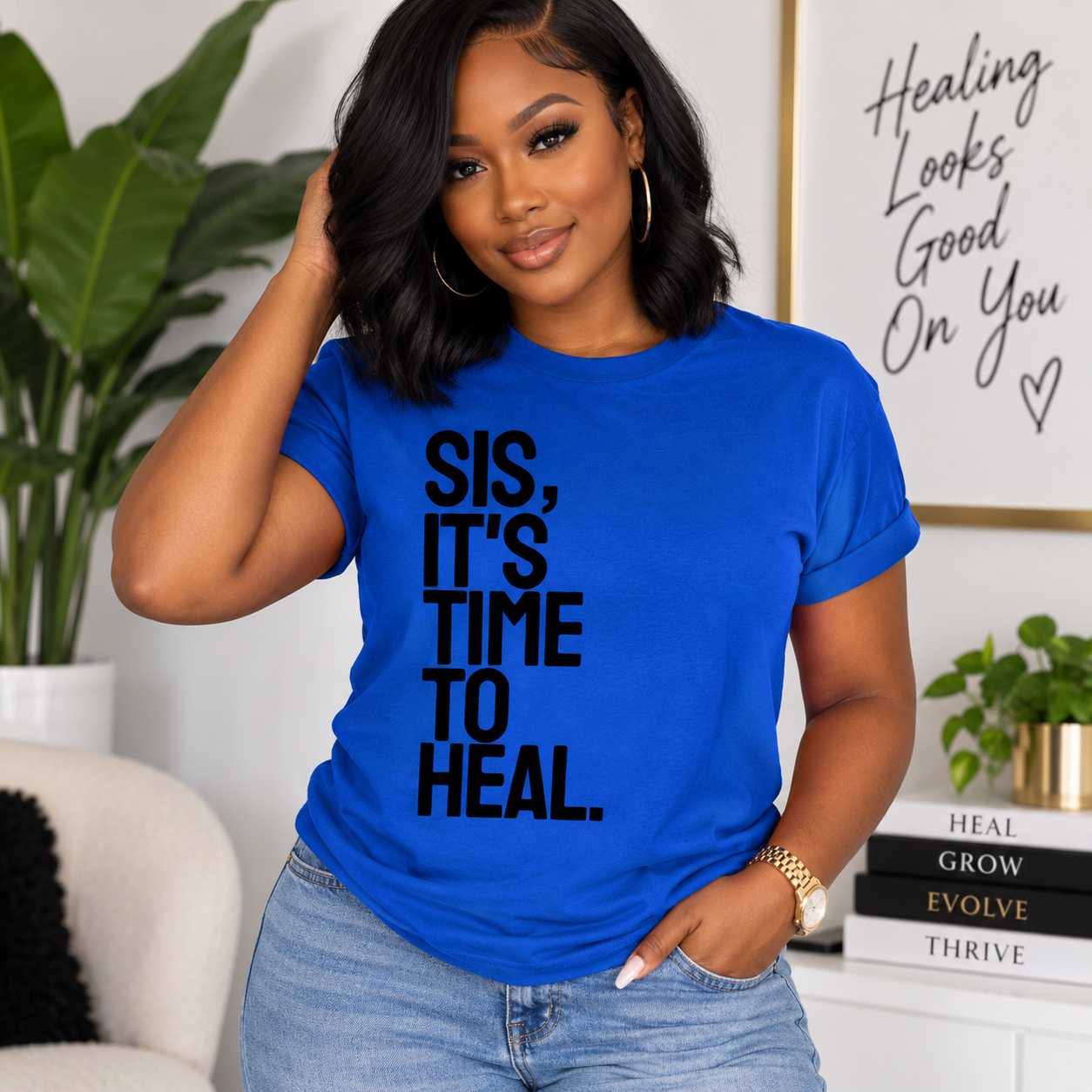 A woman wearing a bright blue graphic t-shirt with the text 'SIS, IT'S TIME TO HEAL.' in bold black letters, paired with light wash blue jeans in a cozy, modern room setting.