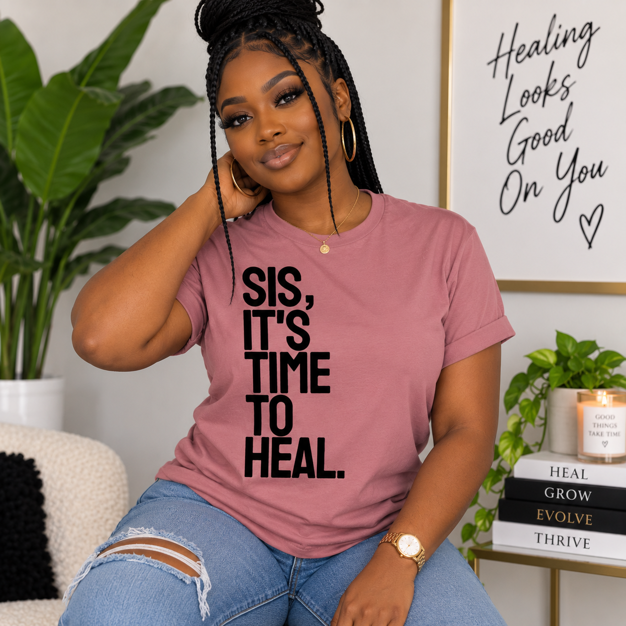 A woman wearing a dusty rose graphic t-shirt with the text 'SIS, IT'S TIME TO HEAL.' in black, paired with distressed blue jeans, sitting in a cozy room setting with plants and home decor.