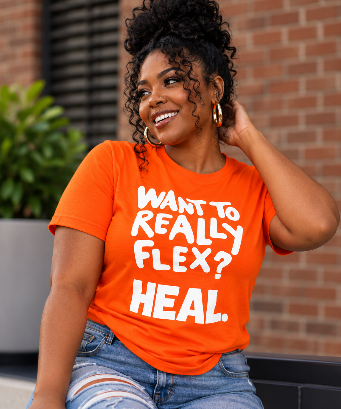 A woman wearing a bright orange graphic t-shirt with the text 'WANT TO REALLY FLEX? HEAL.' in white, paired with distressed blue jeans, posing outdoors against a brick wall background.
