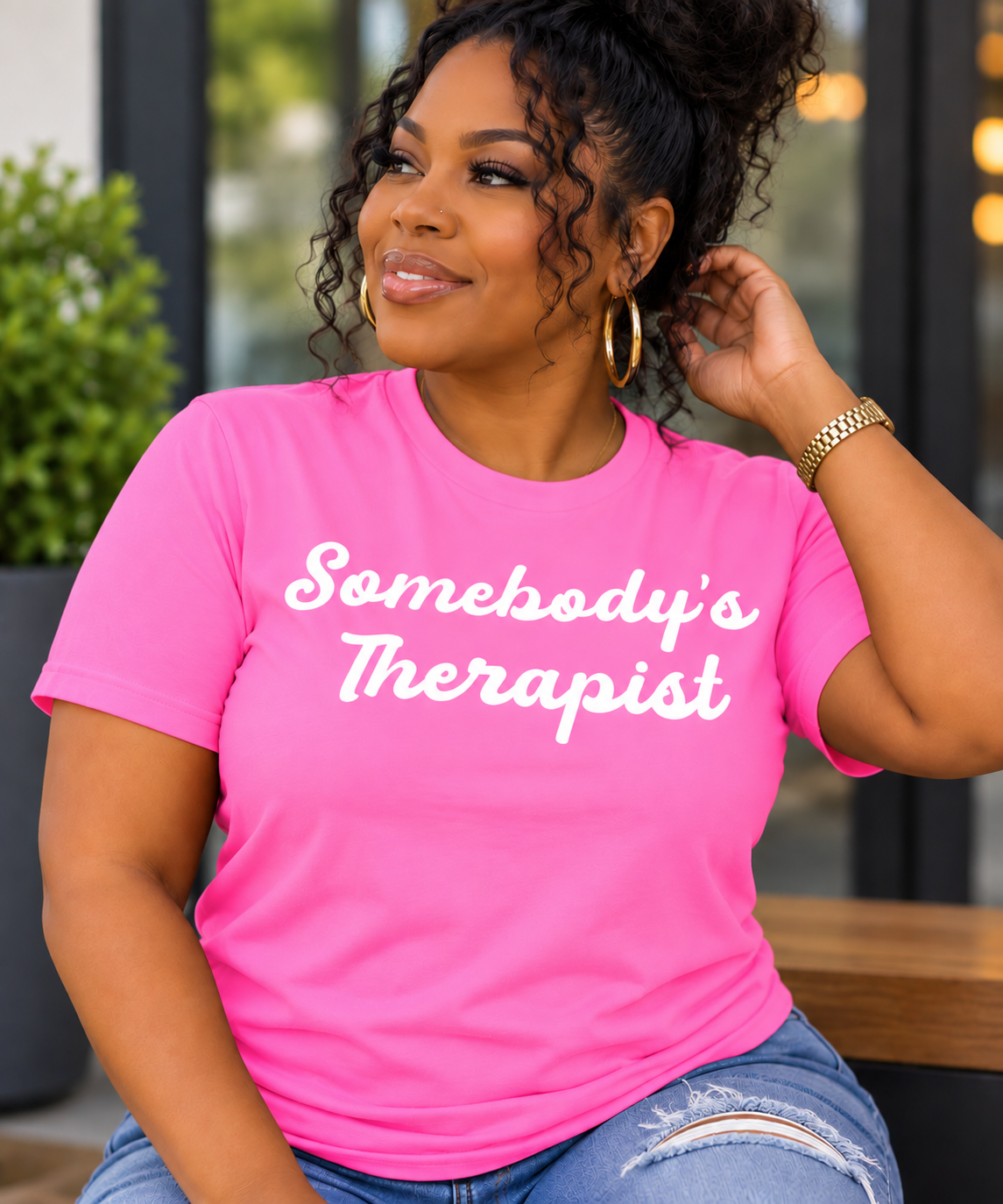 A woman wearing a bright pink t-shirt with the text 'Somebody's Therapist' printed in white script across the chest, paired with blue jeans and gold hoop earrings.