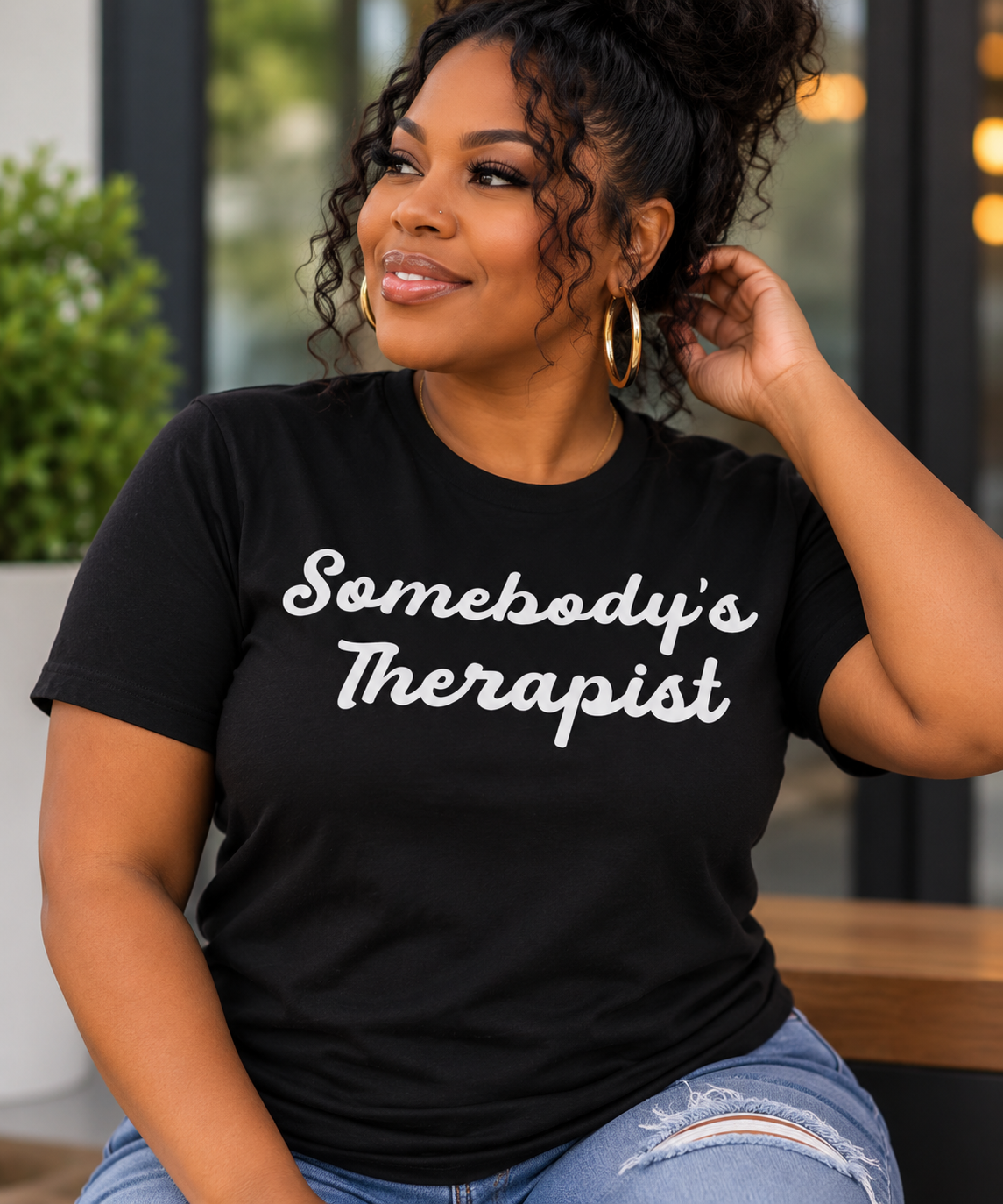 A woman wearing a black graphic t-shirt with the text 'Somebody's Therapist' in white script, paired with light blue distressed jeans, sitting in a modern indoor setting.