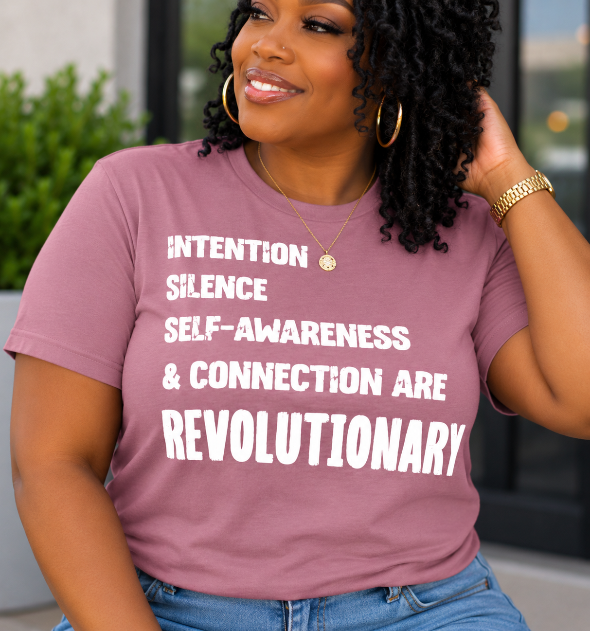A woman wearing a mauve pink graphic t-shirt with bold white text that reads 'INTENTION SILENCE SELF-AWARENESS & CONNECTION ARE REVOLUTIONARY'. She is also wearing blue jeans, gold hoop earrings, and a gold necklace.
