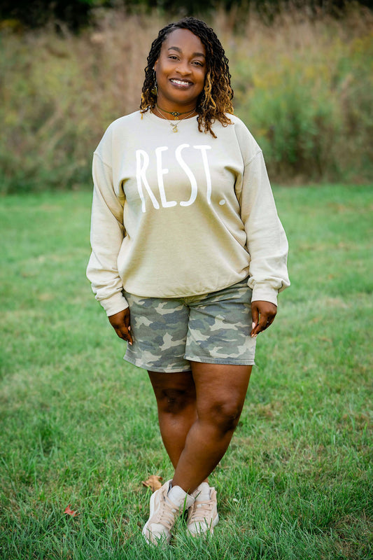 Woman wearing a beige sweatshirt with 'REST' and camouflage shorts standing in a grassy field.